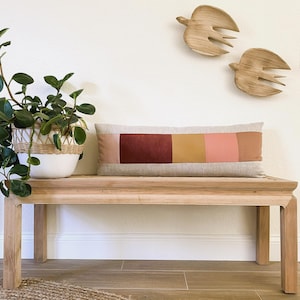 May include: A rectangular cushion with a patchwork design in shades of red, orange, and pink, placed on a light wood bench. A potted plant with green leaves is on the left, and two wooden bird sculptures decorate the wall above.