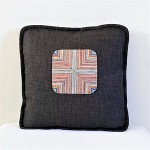 May include: A dark gray throw pillow with a colorful geometric pattern in the center. The pattern is made up of stripes of various colors, including red, orange, yellow, blue, and green.