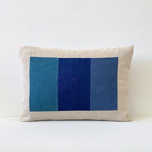 May include: Rectangular decorative pillow with a natural linen border and a central panel of three vertical blue stripes. The stripes vary in shade from teal to navy blue, creating a color block design. The pillow is a neutral home decor item.