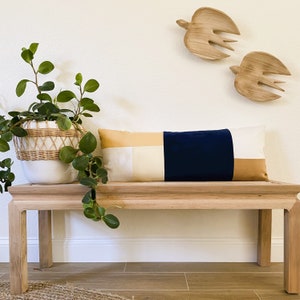May include: A rectangular decorative pillow with a color block design in shades of gold, cream, navy blue, and white. The pillow is placed on a light wood bench. A potted plant and wooden bird sculptures are also visible.