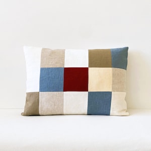 Peut inclure: Un coussin rectangulaire avec un motif patchwork. Le coussin est fait de tissus de différentes couleurs, dont le beige, le blanc, le bleu clair et le rouge foncé. Le coussin est posé sur une surface blanche.