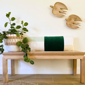 May include: A rectangular decorative pillow with a color block design in shades of green, cream, and white rests on a wooden bench. A potted plant in a woven basket and two wooden bird sculptures complete the decor.