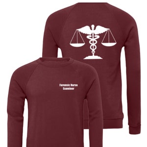 Könnte beinhalten: Burgunderfarbener Sweatshirt mit einem weißen Grafikdruck eines Caduceus und einer Waage auf der Rückseite. Die Vorderseite des Sweatshirts trägt den Text "Forensic Nurse Examiner" in Weiß.