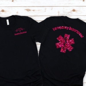 Könnte beinhalten: Schwarzes T-Shirt mit einem rosa Herzen und einer EKG-Linie auf der Vorderseite und dem Text "Emergency Department" sowie einem rosa Glitzer-Stern des Lebens auf der Rückseite.