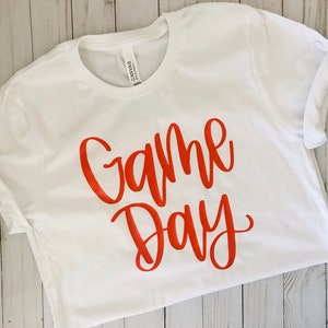 Könnte beinhalten: Weißes T-Shirt mit dem Aufdruck "Game Day" in Orange.