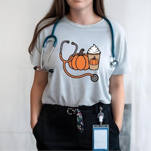 Könnte beinhalten: Ein hellgraues Kurzarm-T-Shirt mit einer Grafik aus einem orangefarbenen Kürbis und einem Kaffeebecher mit Schlagsahne, die durch ein Stethoskop-Design verbunden sind. Ein blaugrünes Stethoskop liegt um den Halsausschnitt.