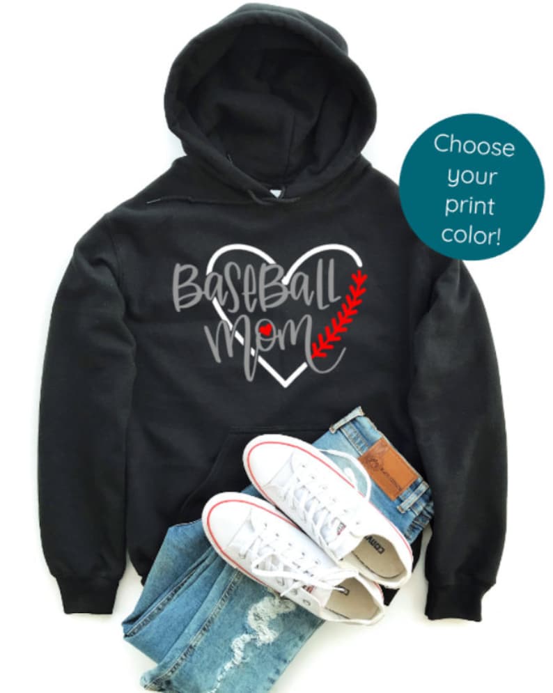 May include: Black hooded sweatshirt with a white heart design that says "Baseball Mom" with a red baseball stitch and a red baseball. The sweatshirt is lying on a pair of blue jeans and white trainers.
