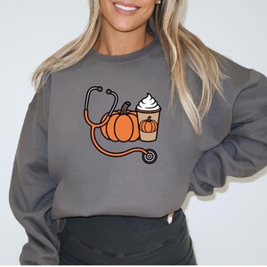 Könnte beinhalten: Ein anthrazitfarbenes Sweatshirt mit Rundhalsausschnitt und einem Grafikdruck, der einen orangefarbenen Kürbis, ein Stethoskop und einen mit Schlagsahne bedeckten Kaffeebecher zeigt. Der Becher hat eine kleine Kürbisabbildung.