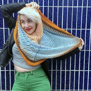 May include: A woman wearing a black leather jacket and green pants is holding a colorful knitted scarf with orange, blue, and white stripes. The scarf is draped over her head and shoulders.