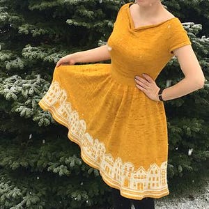 Puede incluir: Vestido amarillo mostaza con un diseño de ciudad blanco y amarillo a lo largo del dobladillo. El vestido tiene mangas cortas y un escote redondo. La persona lleva medias negras y botas negras. El fondo es una escena nevada con un árbol verde.