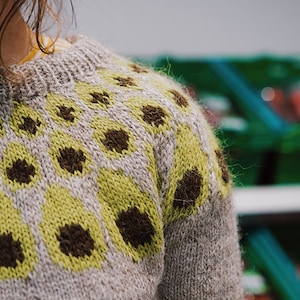 Könnte beinhalten: Nahaufnahme eines grauen Strickpullovers mit einem geometrischen Muster aus grünen und braunen Quadraten.