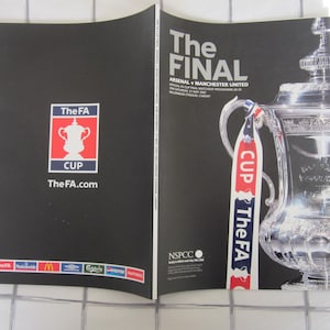 Peut inclure: Programme noir et blanc de la finale de la FA Cup entre Arsenal et Manchester United. Le programme présente le logo de la FA Cup, le texte "The Final" et un trophée argenté. Le programme est ouvert sur une surface à carreaux.