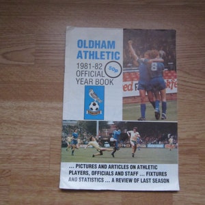 Könnte beinhalten: Ein blauer und weißer Einband für das offizielle Jahrbuch von Oldham Athletic 1981-82. Der Einband zeigt ein Foto von zwei Fußballspielern, die ein Tor feiern. Das Buch kostet 50 Pence.