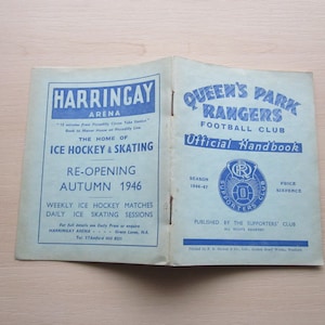 Könnte beinhalten: Eine blaue und weiße Broschüre für den Fußballverein Queen's Park Rangers. Die Broschüre trägt den Titel "Offizielles Handbuch" und zeigt das Logo des Vereins, einen blauen Kreis mit einem weißen "QPR" darin. Die Broschüre ist auf 1946-47 datiert und kostet sechs Pence.