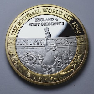 Puede incluir: Moneda conmemorativa con borde dorado y centro plateado. La moneda presenta un jugador de fútbol sosteniendo un trofeo, con el texto "THE FOOTBALL WORLD CUP 1966" y el marcador "ENGLAND 4 WEST GERMANY 2".