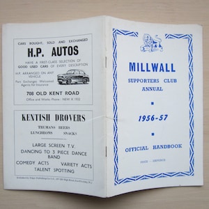 Peut inclure: Un livret noir et blanc avec le titre "Millwall Supporters Club Annual 1956-57" et le texte "Official Handbook Price Sixpence". La couverture avant présente un emblème de lion bleu avec une bordure géométrique bleue et blanche.