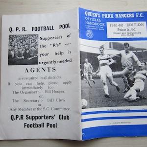 Könnte beinhalten: Ein blaues und weißes Heftchen mit dem Titel "Queen's Park Rangers F.C. Official Handbook 1961-62 Edition". Das Heftchen ist auf einer Seite mit einem Schwarzweißfoto von zwei Fußballspielern in Aktion geöffnet. Der Text "Price 1s. 6d. Edited and Compiled by Bill Clow" ist auf der Seite gedruckt.