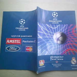 May include: A blue and red program for the UEFA Champions League Final 2002 in Glasgow, Scotland. The program features the UEFA Champions League logo, the official partners, and the teams playing in the final: Bayer Leverkusen and Real Madrid CF. The program also includes the date and time of the match.