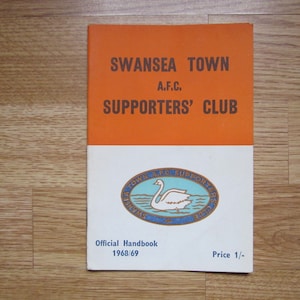 Può includere: Un opuscolo bianco e arancione con il testo "Swansea Town A.F.C. Supporters' Club" e uno stemma con un cigno bianco su sfondo blu. L'opuscolo è intitolato "Official Handbook 1968/69" e ha un prezzo di "1/-."