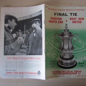 May include: A black and white photo of two men in front of a building with the words "Hot Bovril supporters club" printed below. The image is from a program for the 1962 Football Association Challenge Cup Final between Preston North End and West Ham United. The program cover features a drawing of the FA Cup trophy and the words "Final Tie" and "Wembley".