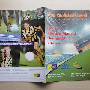 May include: An open magazine featuring soccer players in black and yellow uniforms, with the text "The Gelderland Tournament" and team names. The magazine also includes the text "DE ENERGIE OM TE LEVEN" and the date "4th & 5th of August 1998."