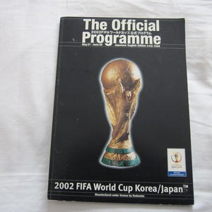 以下が含まれることがあります： 2002 FIFAワールドカップ公式プログラムの黒い表紙。表紙には金色のトロフィーと「The Official Programme」の文字が描かれています。