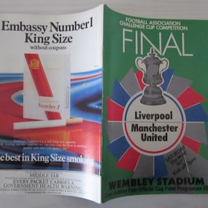 May include: A programme for the 1977 Football Association Challenge Cup Final between Liverpool and Manchester United. The programme features a red, white, and blue starburst design with the FA Cup trophy in the centre. The text "FINAL" is in large, bold letters at the top of the programme. The programme also includes the date of the match, "Saturday 21st May 1977", and the kick-off time, "3pm". The programme is for Wembley Stadium and costs 50p.