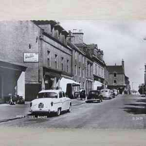 May include: A monochrome postcard depicting High Street, Banff, Scotland, with vintage cars parked along the road. Buildings and shops line the street, and a sign for Walls Ice Cream is visible. The postcard is dated.