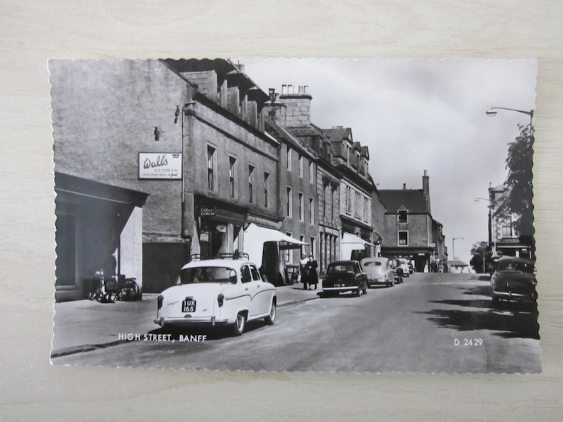 May include: A monochrome postcard depicting High Street, Banff, Scotland, with vintage cars parked along the road. Buildings and shops line the street, and a sign for Walls Ice Cream is visible. The postcard is dated.
