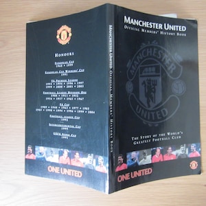 May include: A black and red book titled "Manchester United Official Members' History Book" with a red crest featuring a football and the words "Manchester United". The book cover features a list of the team's honors and a photo of a group of soccer players.