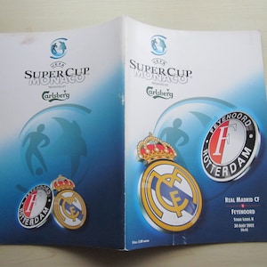 May include: A program for the UEFA Super Cup Monaco match between Real Madrid CF and Feyenoord Rotterdam, held on August 30, 2002. The program features the team logos and the Carlsberg logo.