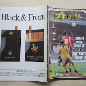 May include: A vintage football match program for the Football Association Challenge Cup Final Replay between Queens Park Rangers and Tottenham Hotspur. The match took place on Thursday, May 27, 1982, at Wembley Stadium. The program features a photo of the players on the field and the text "REPLAY" in large, bold letters.