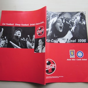 May include: A red and white Coca-Cola Cup Final 1996 program featuring Aston Villa v Leeds United. The program has a black and white photo of a crowd of fans cheering. The program also features the Coca-Cola logo.