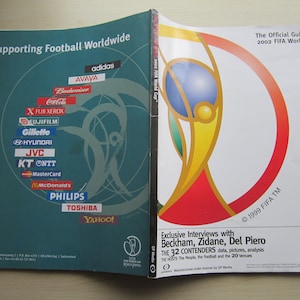 Può includere: Le copertine anteriore e posteriore di una guida per la Coppa del Mondo FIFA 2002. La copertina anteriore presenta i loghi degli sponsor come Adidas, Avaya, Budweiser, Coca-Cola e Gillette. La copertina posteriore presenta il trofeo della Coppa del Mondo FIFA e il testo "The Official Guidebook 2002 FIFA World Cup".