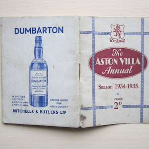 Könnte beinhalten: Ein Vintage-Programm für das Aston Villa Annual, Saison 1934-1935. Das Cover zeigt ein rotes Oval mit weißem Text und einem blau-weiß gestreiften Rand. Der Preis beträgt 2d. Die Rückseite zeigt eine Werbung für Dumbarton Scotch Whisky.