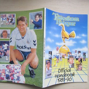 Puede incluir: Un manual oficial de Tottenham Hotspur de la temporada 1989-90, de color blanco y azul. La portada presenta una imagen de dibujos animados de un gallo de pie sobre un balón de fútbol. El manual incluye fotos de jugadores del equipo.