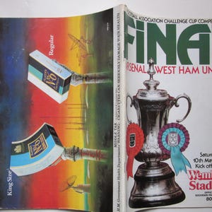 May include: A vintage souvenir program cover for the 1980 Football Association Challenge Cup Final between Arsenal and West Ham United. Features a trophy, ribbons, and the Wembley Stadium logo. Includes cigarette boxes with the text "Players N°6".