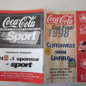Könnte beinhalten: Ein Coca-Cola-Programm für das Finale des Cliftonville gegen Linfield Cup 1998. Das Programm zeigt das Coca-Cola-Logo, die Logos der Teams sowie Datum und Uhrzeit des Spiels. Das Programm ist rot, weiß und blau.