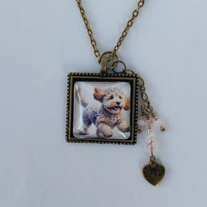 May include: A bronze necklace with a square pendant featuring a picture of a small, white dog with brown markings. The pendant has a beaded charm with a heart-shaped charm that says "love".
