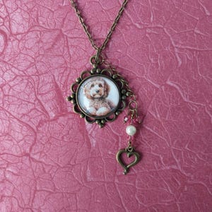 May include: A bronze necklace with a round pendant featuring a brown and white dog. The pendant is surrounded by a decorative frame and has a small heart charm hanging from it.