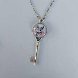 May include: Silver-toned key pendant necklace. The pendant features a round glass cabochon with a watercolor image of a gray kitten surrounded by colorful flowers. The key has a decorative handle and a chain.