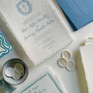 Puede incluir: Juego de invitaciones de boda con papel texturizado y texto verde y azul verdoso. Incluye tarjetas de invitación, tarjetas RSVP y sellos de cera. La invitación principal tiene un escudo floral con las iniciales "BZ".