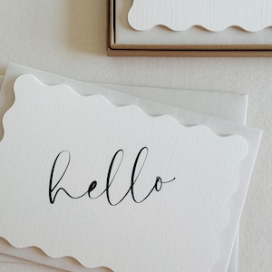 Könnte beinhalten: Weiße Schreibwarenkarten mit wellenförmigem Rand und dem Wort „hello“ in schwarzer Schrift. Die Karten liegen auf einem weißen Umschlag. Eine ähnliche Karte ist in einer braunen Schachtel im Hintergrund zu sehen. Das Schreibwarenset liegt auf einer weißen Oberfläche.