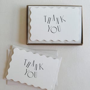 Puede incluir: Un conjunto de tarjetas de agradecimiento blancas con borde festoneado y texto negro, presentadas en una caja marrón. Se ven tarjetas adicionales fuera de la caja. Las tarjetas tienen un diseño sencillo y elegante.