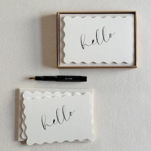 Puede incluir: Un juego de tarjetas rectangulares de color blanco roto con bordes festoneados, cada una con la palabra hello en letra cursiva negra. Las tarjetas se presentan en una caja dorada junto a una pluma estilográfica negra con punta dorada.