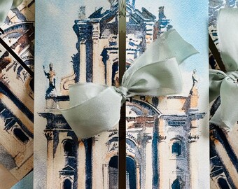 May include: Watercolor-style invitation cards featuring a detailed illustration of a building with columns and a light blue sky. Each card is tied with a light green satin ribbon. The cards are arranged in a stack, with some visible in the background.