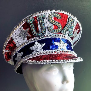May include: A red, white, and blue sequined hat with the word "USA" in rhinestones. The hat has a brim and a pointed top. The hat is decorated with silver stars and rhinestones.