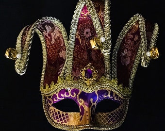 Könnte beinhalten: Eine lila und goldene Narrenmaske mit einem spitzen Hut und dekorativen Details. Die Maske hat ein traditionelles Narren-Design mit einem breiten, offenen Mund und einer kleinen, spitzen Nase.