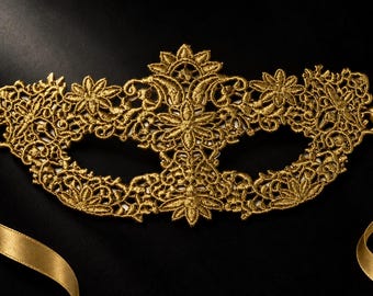May include: A gold lace masquerade mask featuring an intricate floral and scrollwork embroidery pattern. The mask has two eye openings and is attached to two long, smooth gold satin ribbons on each side for securing the item in place.