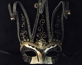 Könnte beinhalten: Eine schwarze und goldene Narrenmaske mit weißem Gesicht und filigranen Details. Die Maske hat drei Spitzen oben und Glocken an den Seiten.